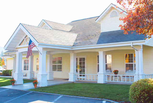 Front exterior of a single-story senior living building with a covered porch, white columns, an American flag, and a manicured lawn.