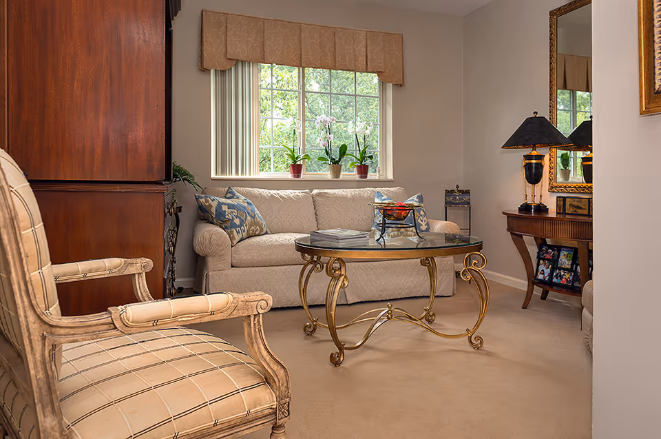 Cozy living room with a sofa, patterned armchair, glass-top coffee table, window with potted plants, and a side table with a lamp.