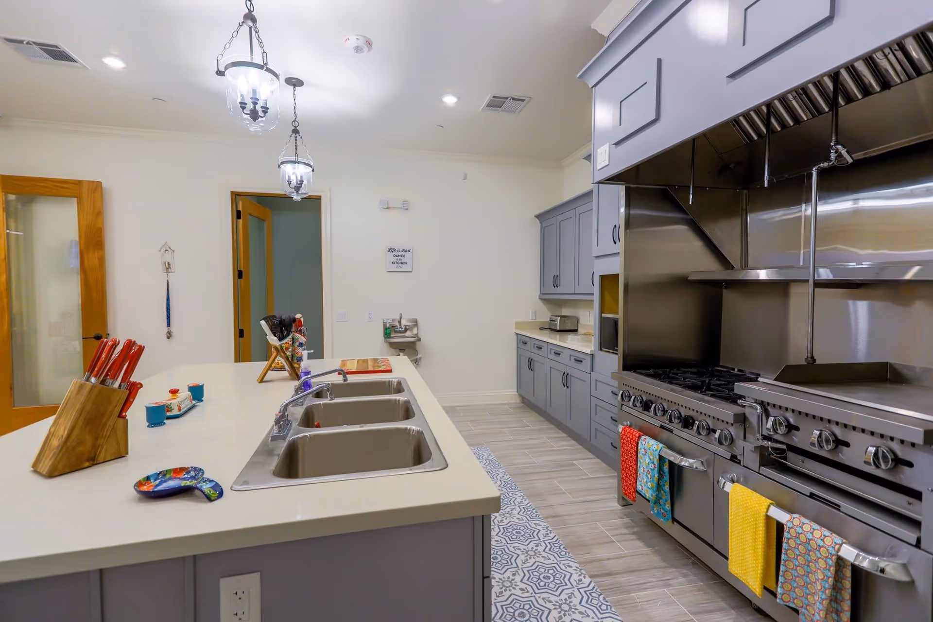 A modern kitchen with a large island featuring three stainless steel sinks and a set of red-handled knives. The kitchen has gray cabinets, a large stainless steel stove with multiple burners and ovens, and colorful towels hanging on the oven handles. Two pendant lights hang from the ceiling, and there is a wooden door with glass panels in the background.