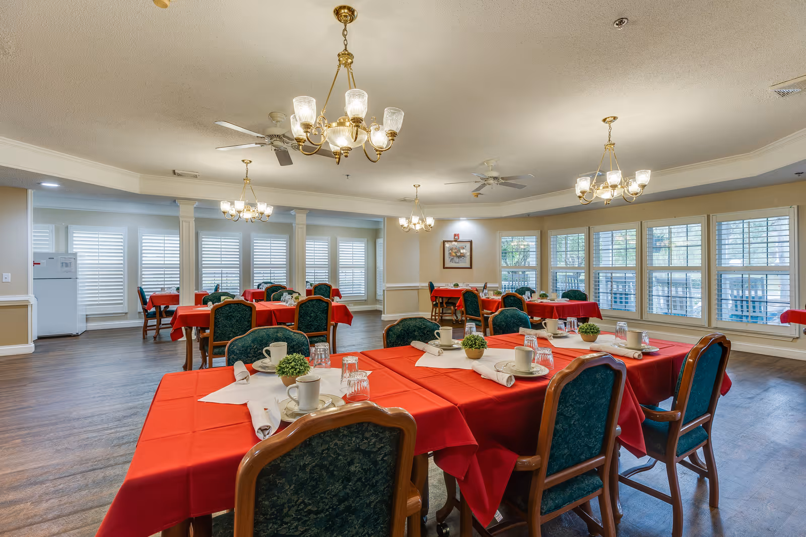 A dining room with multiple tables covered in red tablecloths, each set with white napkins, cups, glasses, and small potted plants. The room has large windows with white shutters, wooden flooring, and several chandeliers hanging from the ceiling. Green cushioned chairs surround the tables.
