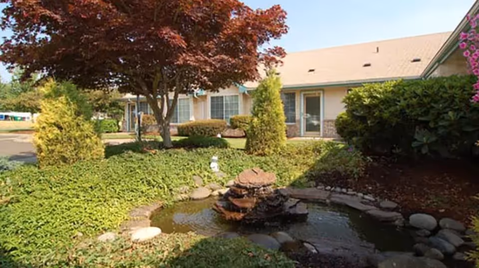Landscaped courtyard with a small pond and fountain in front of a single-story senior living building with trees and shrubs.