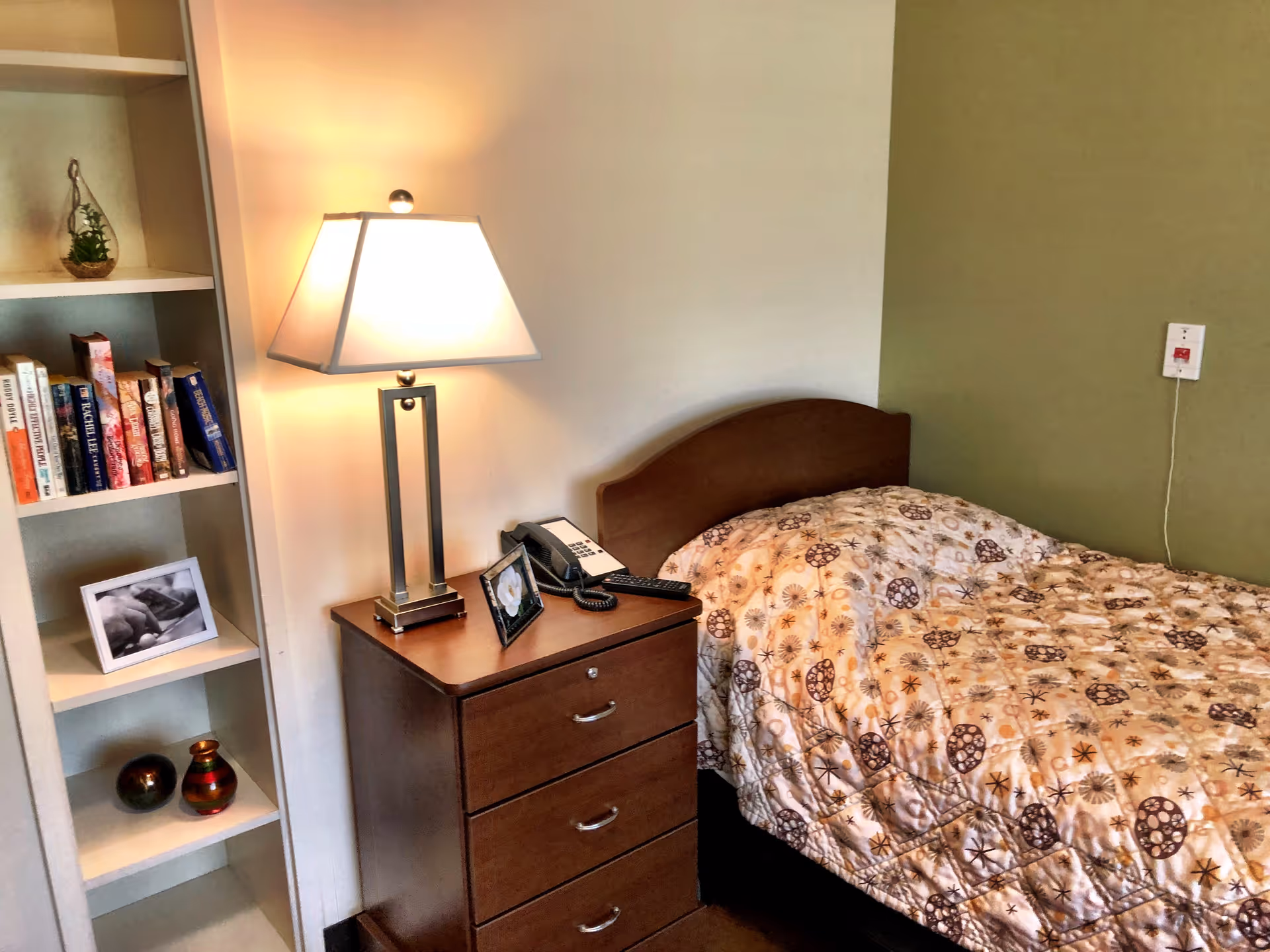 A cozy bedroom corner in a senior living facility featuring a single bed with a patterned quilt, a wooden nightstand with a lamp, telephone, and framed photo, and a built-in bookshelf with books, a framed black and white photo, and decorative items.