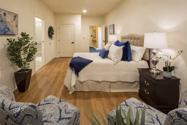 A well-lit bedroom in a memory care community featuring a neatly made bed with beige bedding and blue accent pillows. There are two patterned armchairs in the foreground, a dark wooden nightstand with a lamp and a small plant, and a potted plant near the wall. The room has wooden flooring and light-colored walls, with a door leading to a bathroom and a hallway visible in the background.