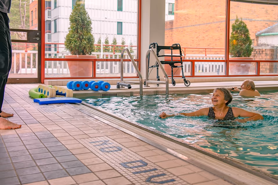 An indoor swimming pool area with two elderly people enjoying the water. One woman is smiling and swimming near the edge, while an elderly man is in the background. Pool equipment such as foam dumbbells and a walker are visible near the poolside. Large windows show an outdoor view with buildings and potted plants.