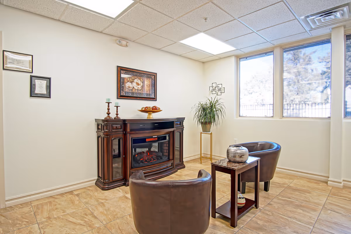 Small furnished lounge with two leather chairs, a side table, decorative fireplace cabinet, wall art, and windows.