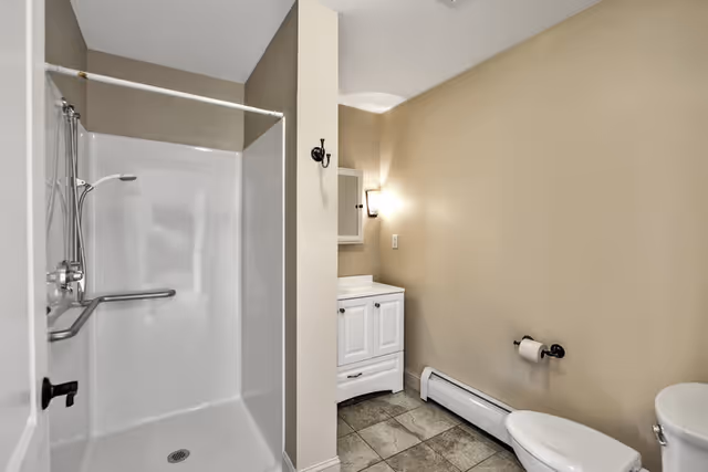 A clean bathroom featuring a white shower stall with a grab bar and handheld showerhead, a white vanity with a mirror and wall-mounted light above it, a toilet with a toilet paper holder on the wall, beige walls, and tiled floor.