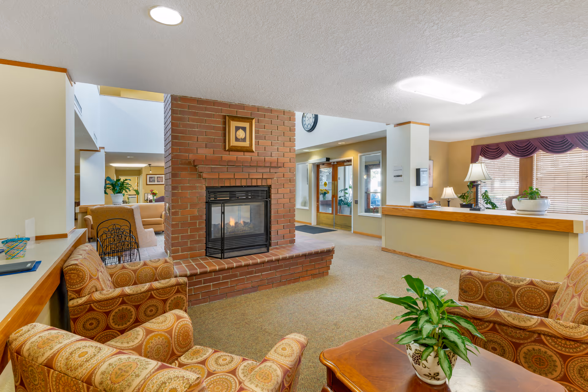 A cozy senior living facility common area with a brick fireplace in the center, surrounded by patterned armchairs and a wooden coffee table with a potted plant. The room has beige walls, carpeted floors, and large windows with blinds and a purple valance. A reception desk with lamps and plants is visible to the right, and double glass doors lead outside in the background.