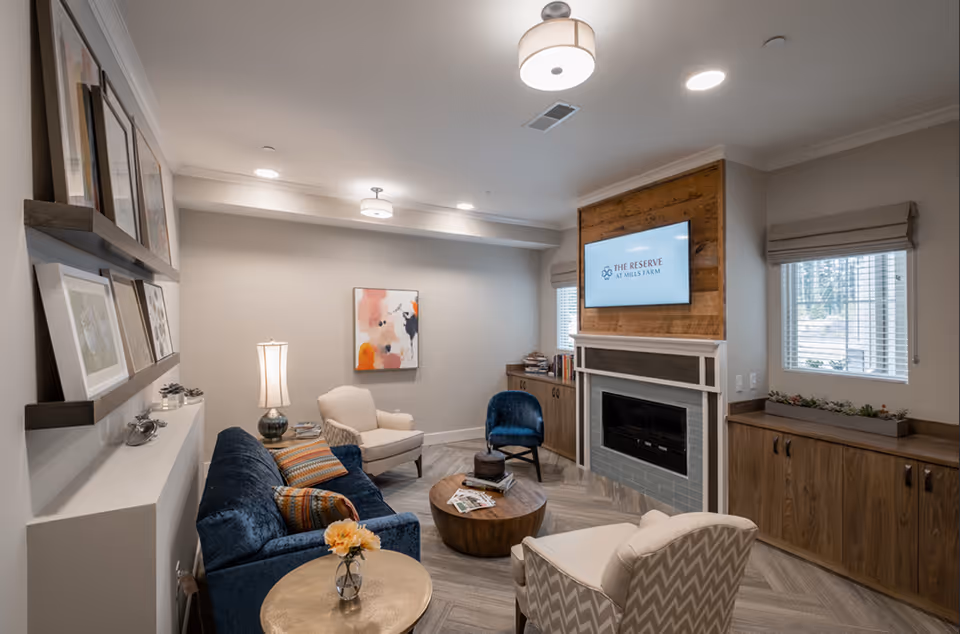 A cozy living room with a blue velvet sofa, two white armchairs, a round wooden coffee table, and a side table with a flower vase. The room features a fireplace with a wooden accent wall above it, where a TV screen displays 'The Reserve at Mills Farm'. There are two windows with blinds, built-in wooden cabinets, and framed artwork on the walls.