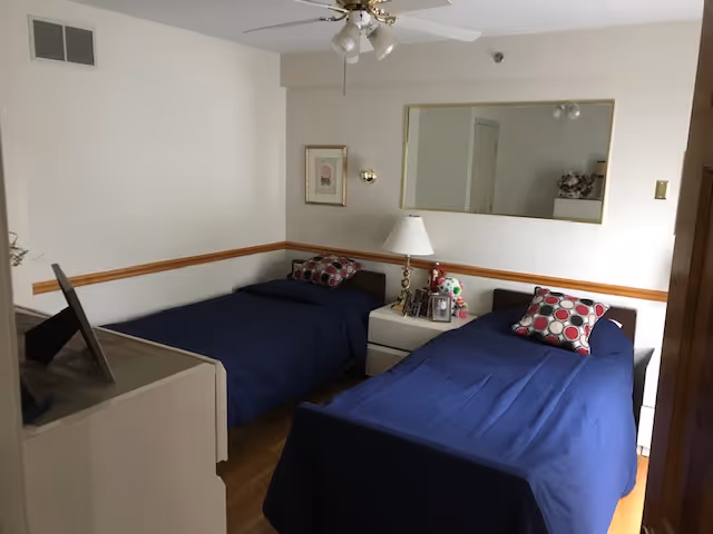 A small bedroom with two single beds covered in dark blue bedspreads, each with a decorative pillow featuring a red, white, and black circular pattern. Between the beds is a white nightstand with a lamp, framed photos, and a small stuffed animal. A large rectangular mirror hangs on the wall above the nightstand, and a ceiling fan with lights is mounted on the ceiling. The room has light-colored walls with a wooden chair rail and hardwood flooring.