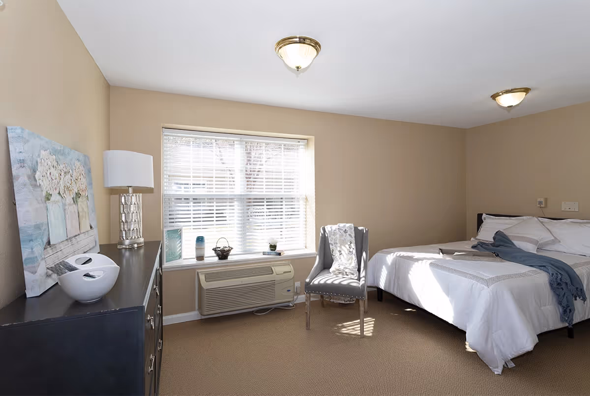 A bright bedroom with beige walls and carpet, featuring a neatly made bed with white bedding and a blue throw blanket. There is a gray upholstered chair with a patterned throw draped over it near a window with white blinds. A black dresser with decorative items and a lamp is positioned against one wall.