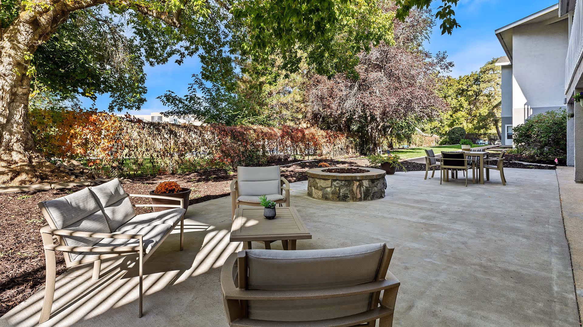 Outdoor patio area at Harrison Regent Retirement Community with cushioned seating, a stone fire pit, and a dining table with chairs. The patio is surrounded by trees and greenery under a clear blue sky.