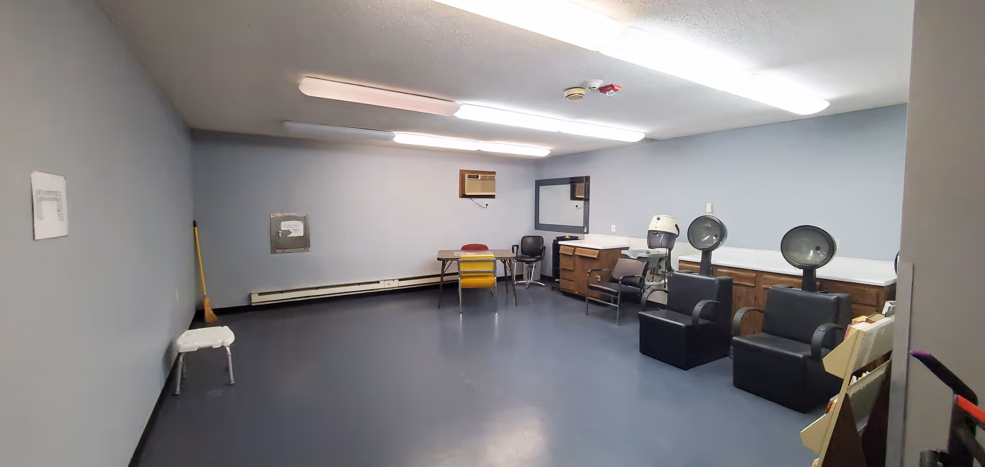 Large interior room with salon hairdryers, chairs, a table and cabinets under fluorescent lights.