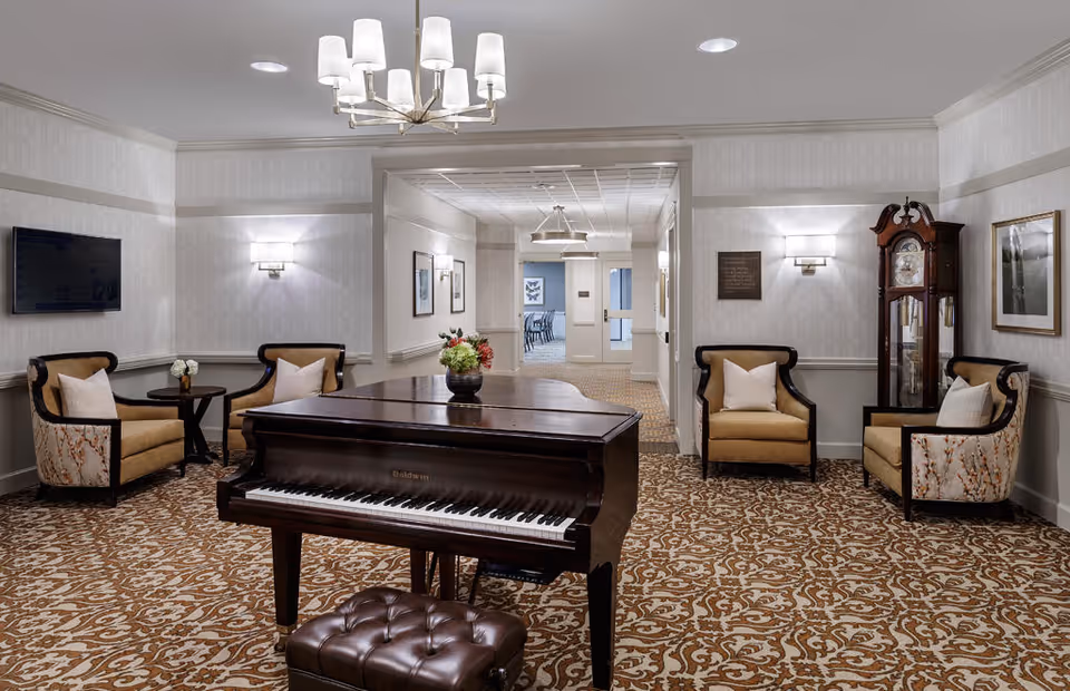 A cozy interior room featuring a grand piano with a floral arrangement on top, surrounded by four upholstered armchairs with pillows. The room has patterned carpet, soft wall lighting, a wall-mounted TV, framed artwork, and a tall grandfather clock. The space opens into a hallway leading to another room with tables and chairs.