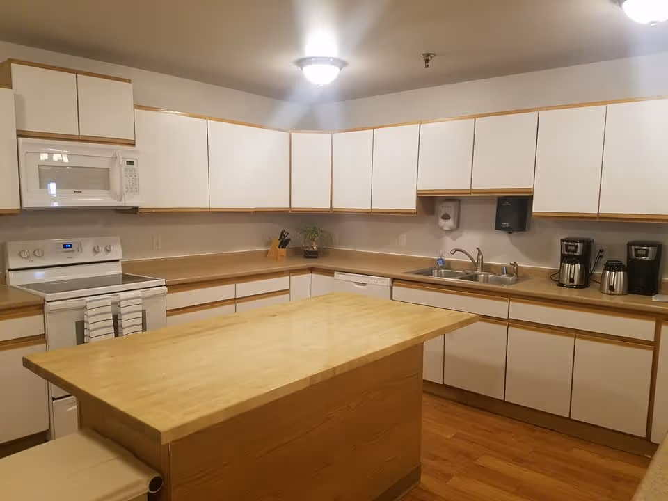 A clean, well-lit kitchen with white cabinets and wood trim. The kitchen features a central wooden island, an electric stove with a microwave above it, a double sink, a dishwasher, and several coffee makers on the countertop. The floor is wooden, and the ceiling has a light fixture.
