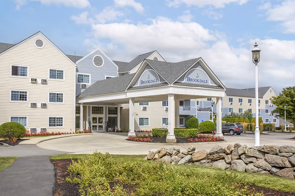 Exterior view of Brookdale Sakonnet Bay senior living facility showing a large entrance canopy with the Brookdale logo, surrounded by landscaped greenery, a stone wall, and a parking area with cars under a partly cloudy sky.