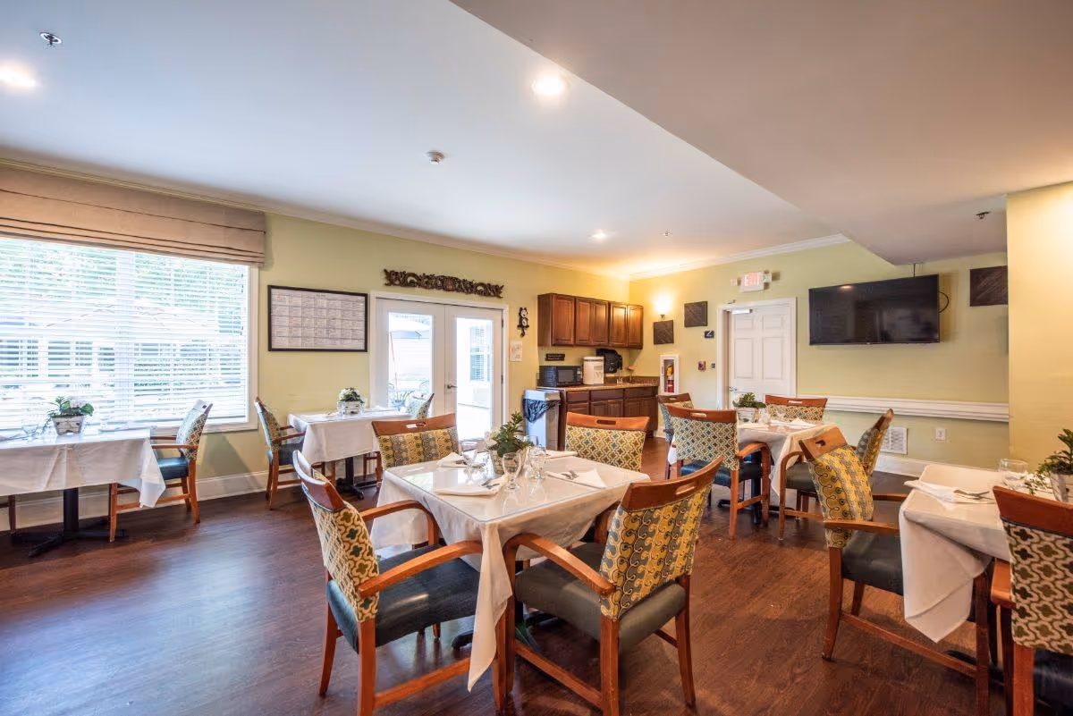 Bright dining room with several tables set with white tablecloths and patterned chairs, large windows on the left and a wall-mounted TV on the right.