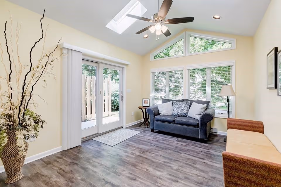 A bright living room with light yellow walls, a ceiling fan with lights, and a skylight. The room features a dark blue loveseat with three decorative pillows, a small wooden side table with a framed photo, a floor lamp, and a cushioned bench with orange and beige upholstery. Large windows with white blinds and sliding glass doors provide a view of greenery outside. A tall vase with decorative branches is placed in the corner.