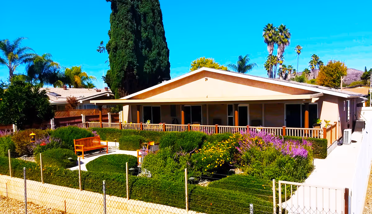 Single-story building with a covered porch surrounded by a well-maintained garden featuring green hedges, colorful flowers, and a wooden bench under a clear blue sky.