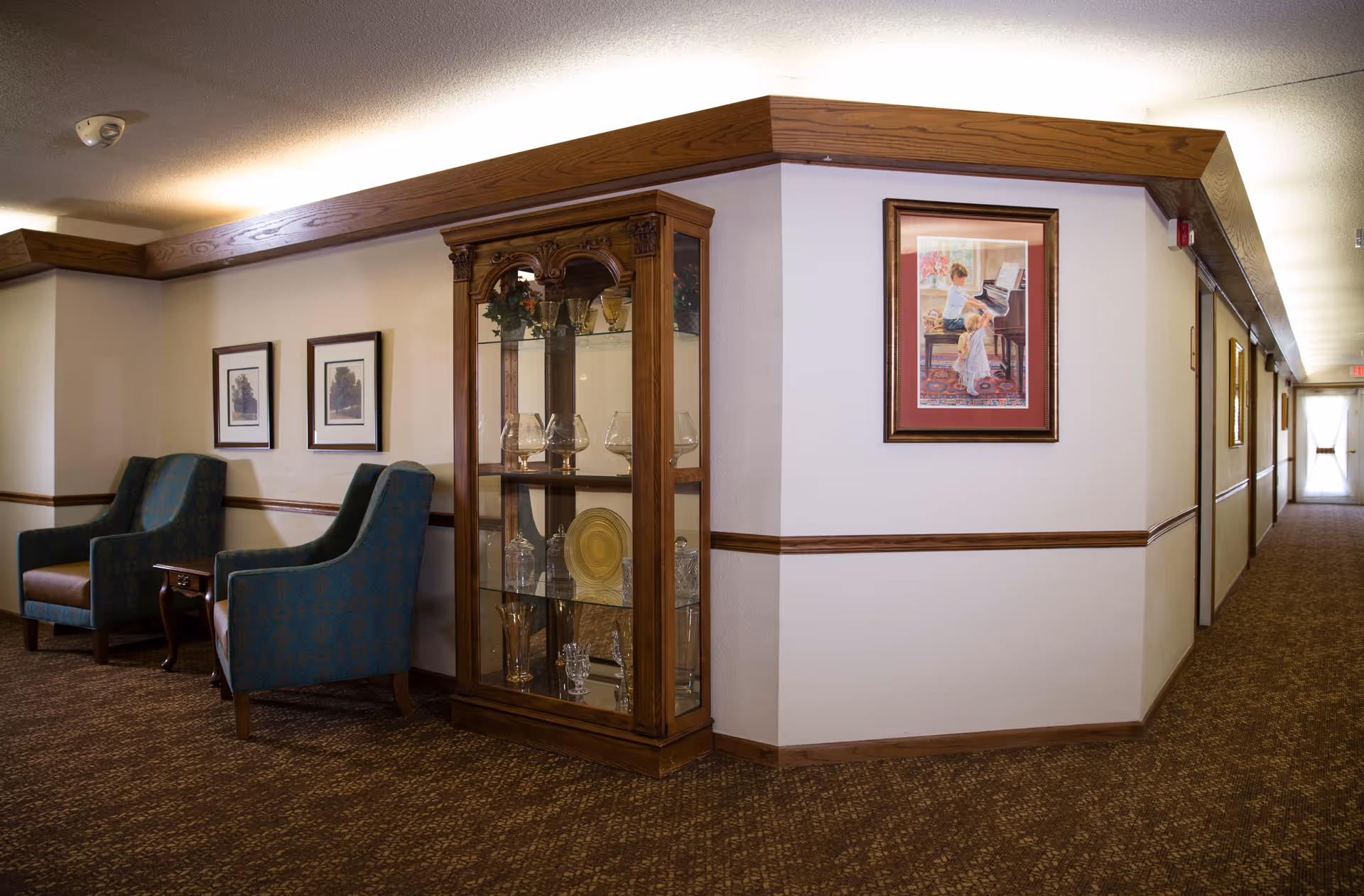 Carpeted interior hallway and sitting area with two upholstered chairs, a glass display cabinet of china, framed artwork, and a long corridor.