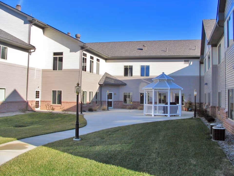 Outdoor courtyard area of a retirement community with a white gazebo in the center, surrounded by a paved walkway, green grass, and a two-story building with beige and brown siding under a clear blue sky.