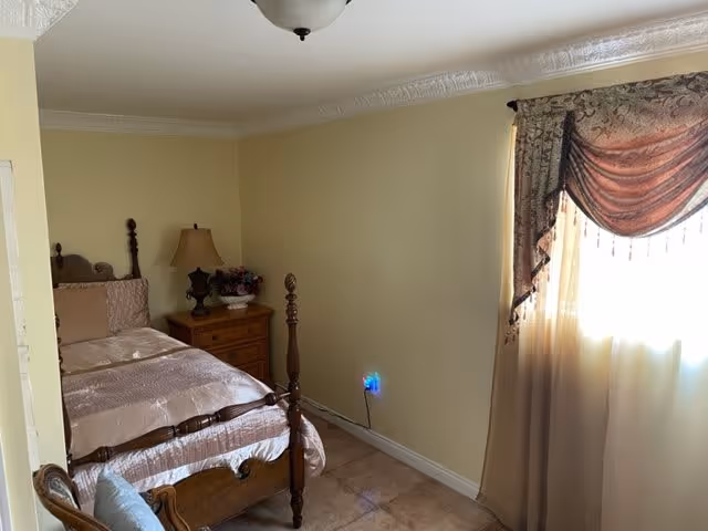 A bedroom with a wooden four-poster bed covered with a light pink satin bedspread. Next to the bed is a wooden nightstand with a lamp and a decorative flower arrangement. The room has beige walls, tiled floor, and a window with sheer curtains and an ornate valance allowing sunlight to enter.