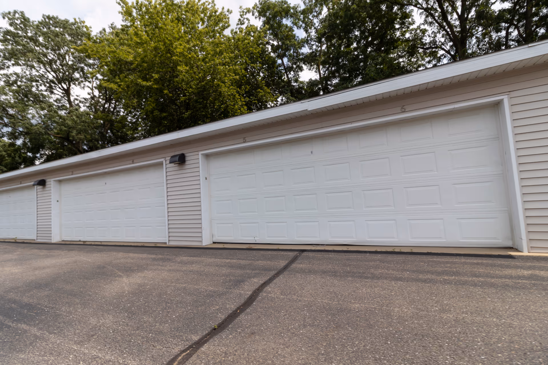 Row of white garage doors on a beige building exterior with trees overhead and a paved driveway in front.