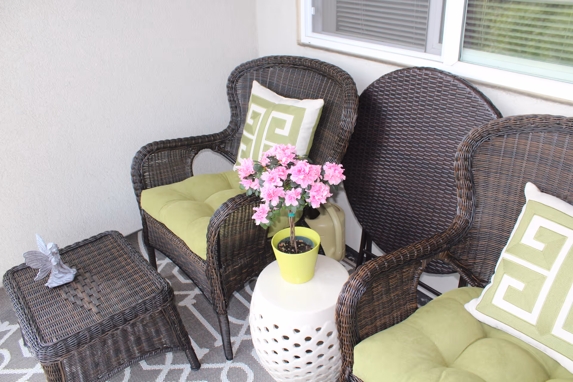 A cozy outdoor seating area with two wicker chairs featuring green cushions and patterned pillows, a small wicker table with a butterfly decoration, and a white ceramic side table holding a potted plant with pink flowers. The area is set against a light-colored wall with a window above.