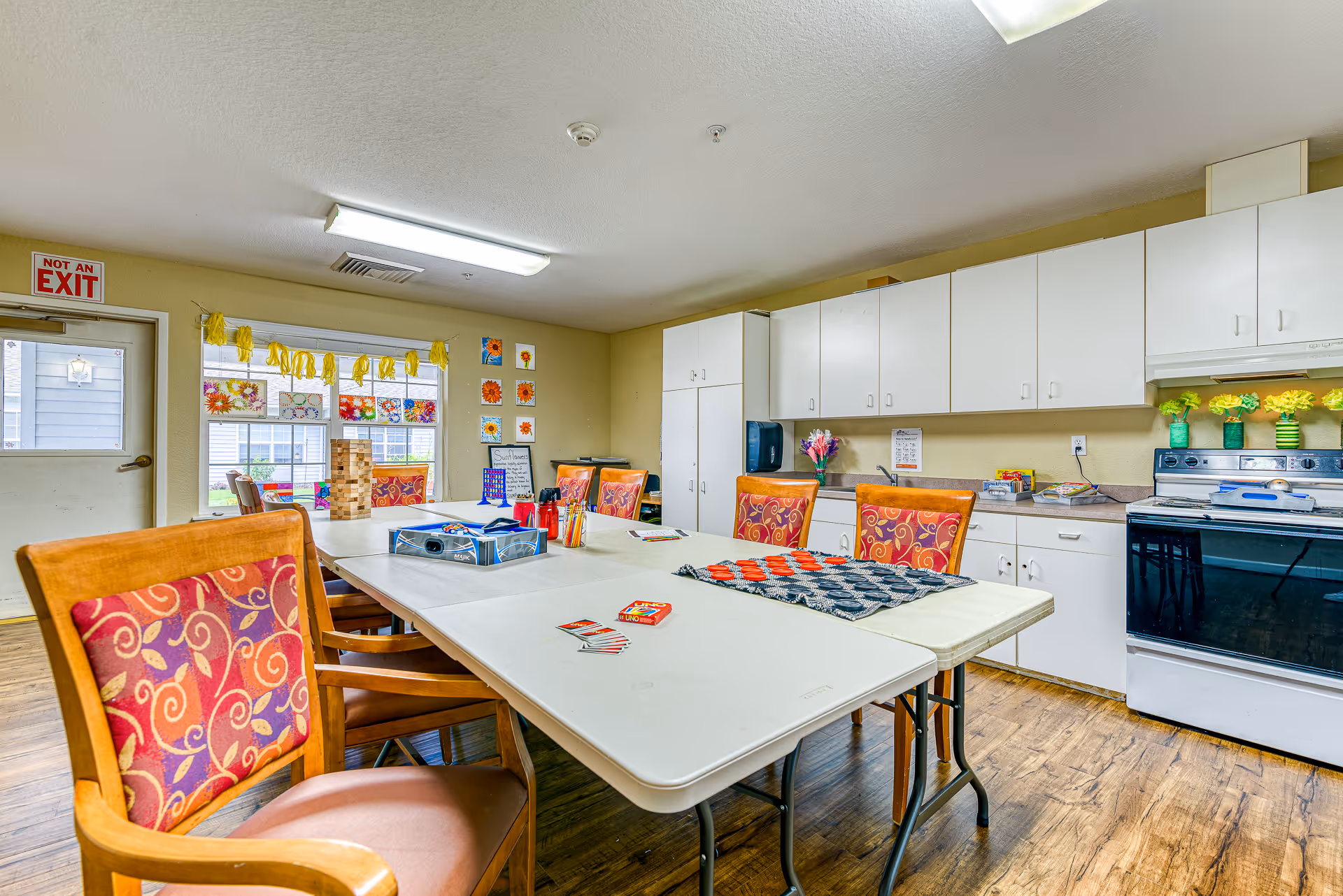 A bright activity room in Creekside Place Memory Care featuring a long table with chairs around it. The table has board games, cards, and craft supplies. The room has white cabinets, a stove, and a countertop with decorative flowers. Windows with colorful artwork and yellow decorations hang on the wall. A door with a 'Not an Exit' sign is visible.