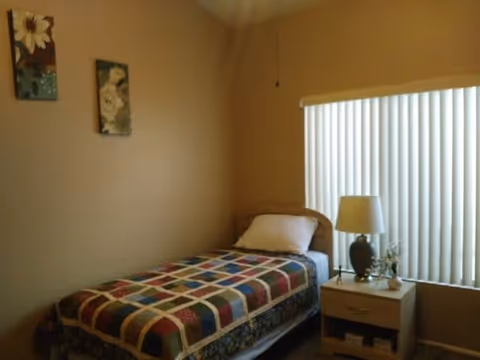 A small bedroom with a single bed covered with a colorful patchwork quilt. There is a white pillow on the bed. Next to the bed is a wooden nightstand with a dark lamp, a small decorative item, and a window with vertical blinds partially closed. Two floral paintings hang on the beige wall above the bed.