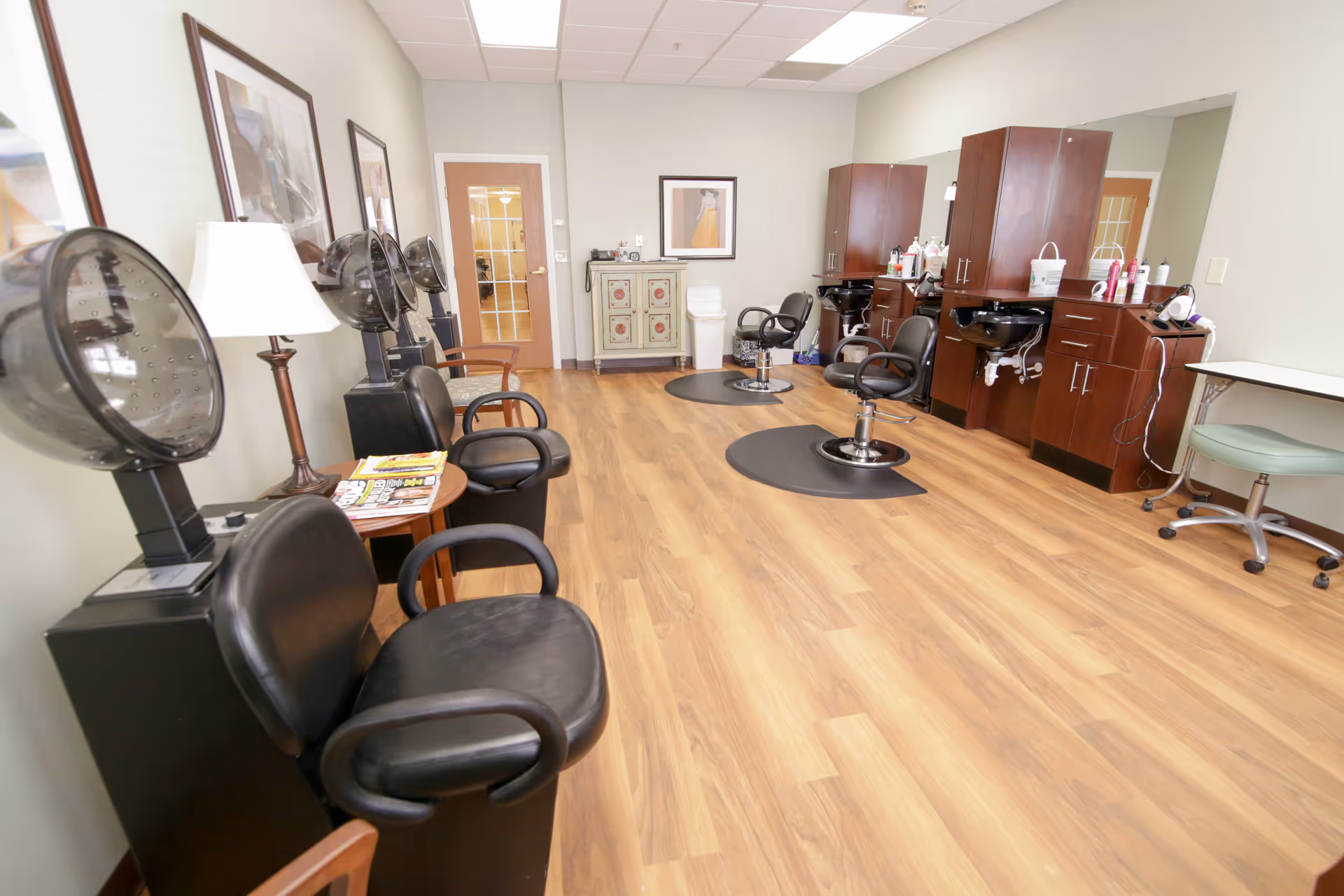 Interior view of a hair salon area with wooden flooring, several black salon chairs, hair dryers, and wooden cabinets with sinks. There are framed pictures on the walls and a small table with magazines and a lamp.