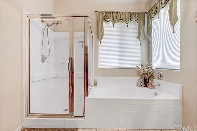 A bathroom with a glass-enclosed shower and a large soaking tub beneath two curtained windows.