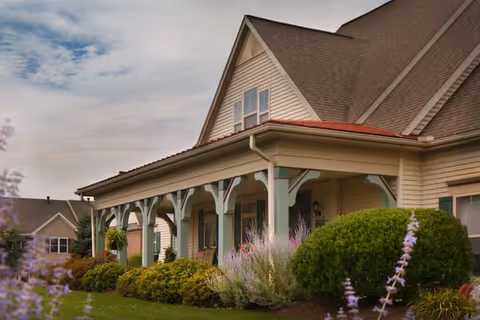 Exterior view of a beige two-story house with a large covered porch supported by teal and white columns, surrounded by well-maintained bushes and flowering plants under a partly cloudy sky.