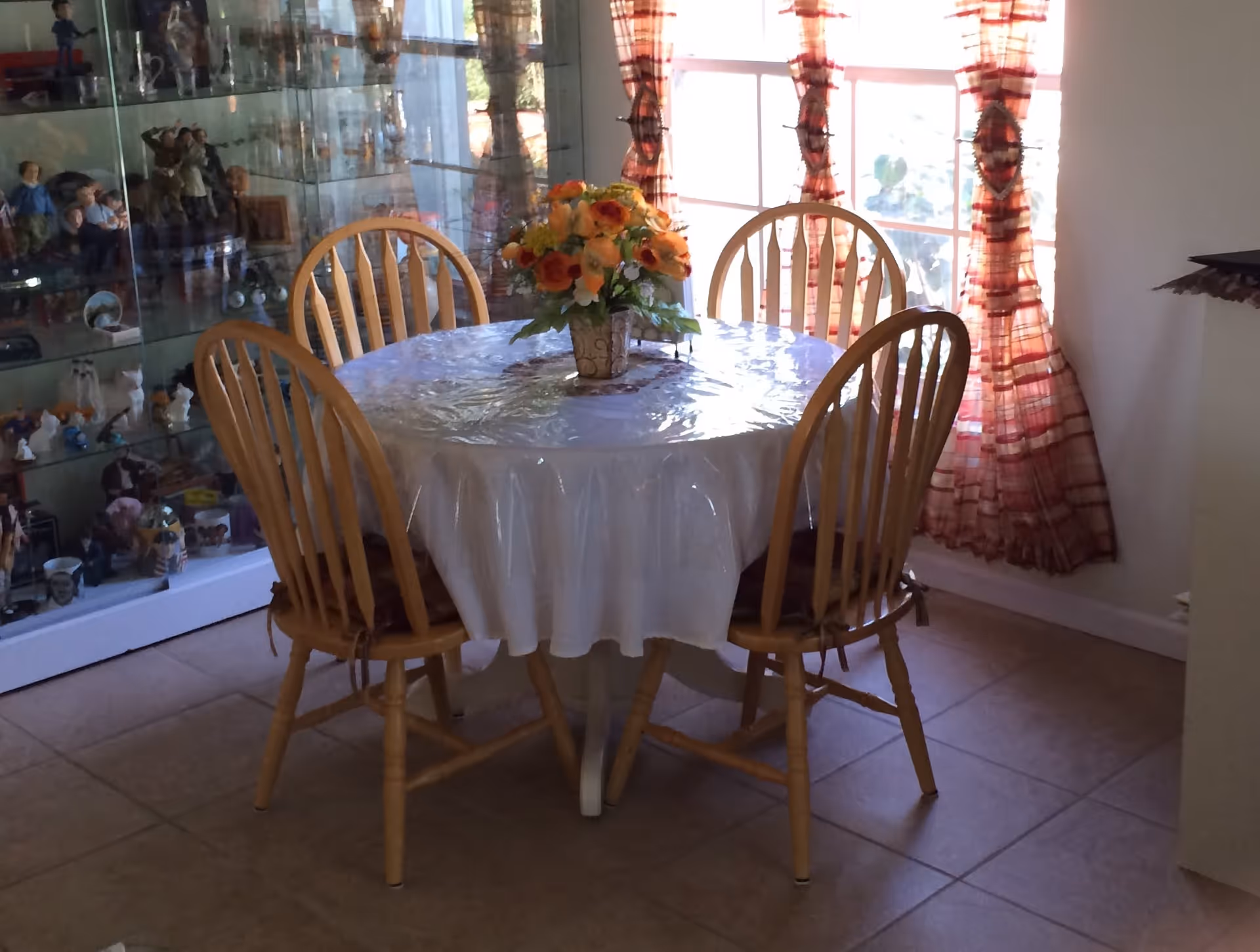 Round dining table with four wooden chairs and a floral centerpiece in a sunlit room with a display cabinet and window curtains.
