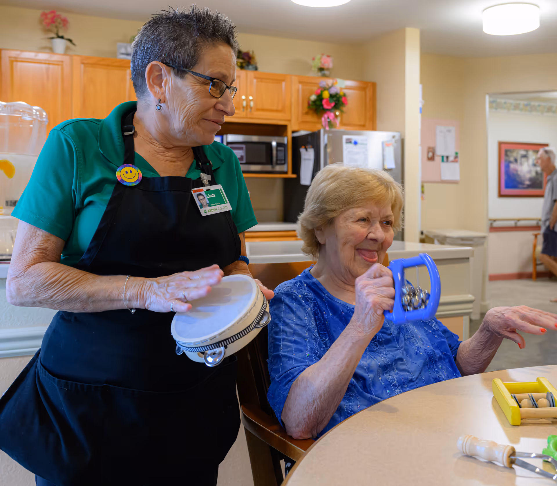 An elderly woman sitting at a table playing with a blue tambourine-like instrument, smiling and engaged in music activity with a staff member wearing a green shirt and black apron who is playing a small drum in a kitchen area.