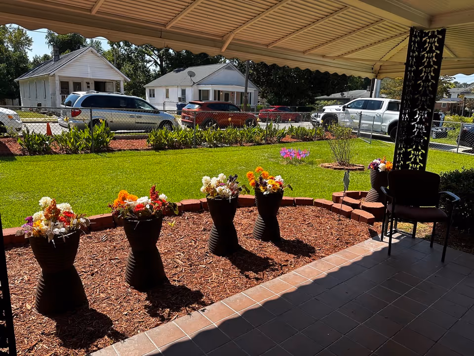 Covered patio with potted flowers and chairs overlooking a grassy yard and houses across the street.