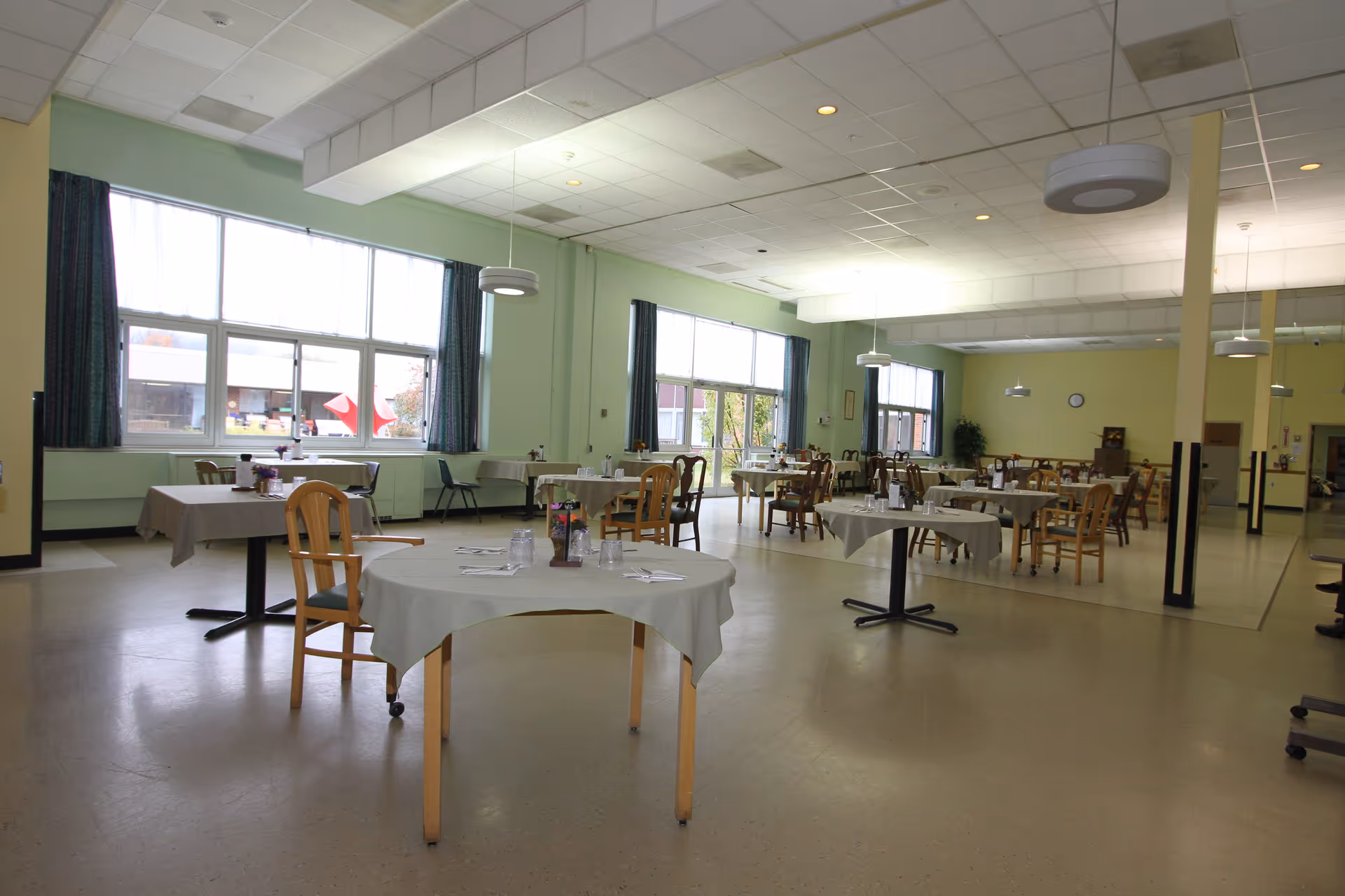 A spacious dining room with multiple round tables covered with white tablecloths, each set with glasses, napkins, and utensils. Wooden chairs are arranged around the tables. Large windows with curtains allow natural light to fill the room. The walls are painted light green and the ceiling has recessed lighting and hanging fixtures.