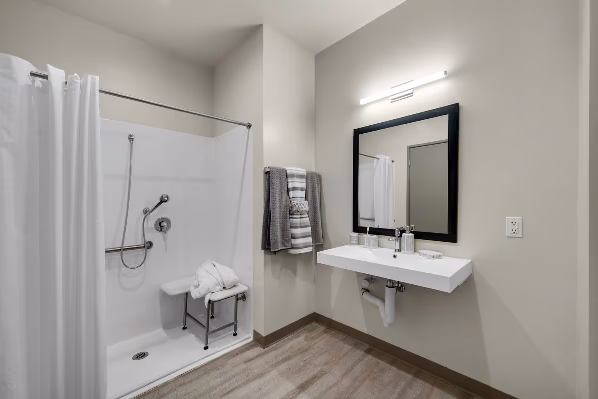 A clean and modern bathroom with a walk-in shower featuring a white curtain, a shower seat with a folded towel on it, and a handheld showerhead. There is a white floating sink with a black-framed mirror above it, a light fixture above the mirror, and a towel rack with neatly folded towels on the wall.