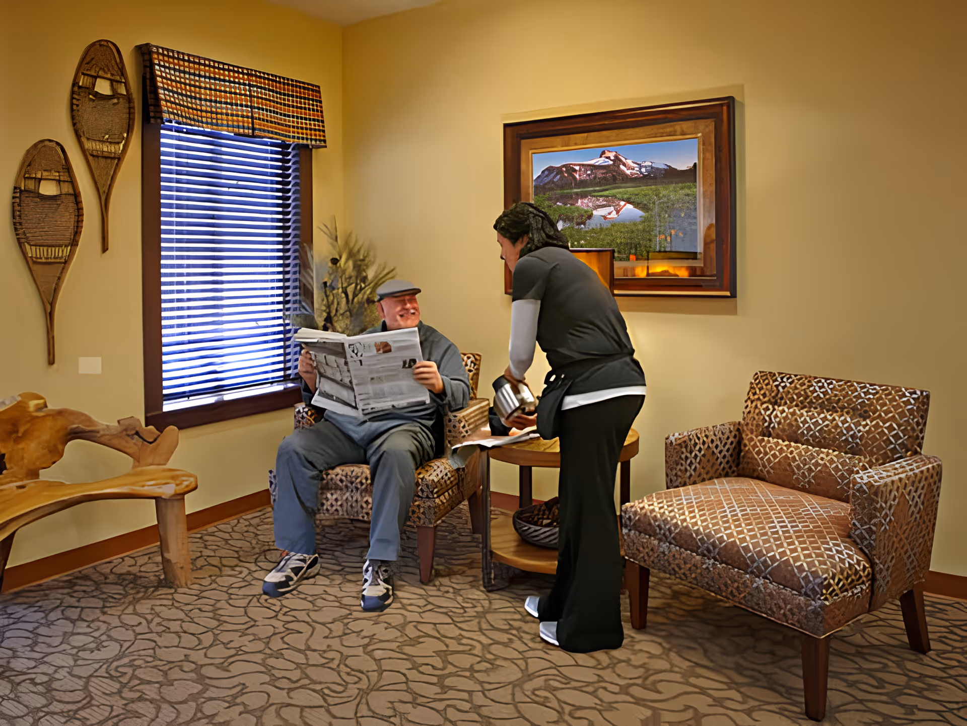 A senior man sitting in a patterned armchair reading a newspaper while a caregiver pours a drink for him in a cozy room with a window covered by blinds and a valance, wooden snowshoe decorations on the wall, a wooden bench, and a framed landscape picture.