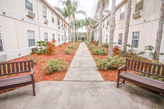 A central paved walkway lined with benches, landscaped beds and palm trees between two three-story buildings leading to a small gazebo.