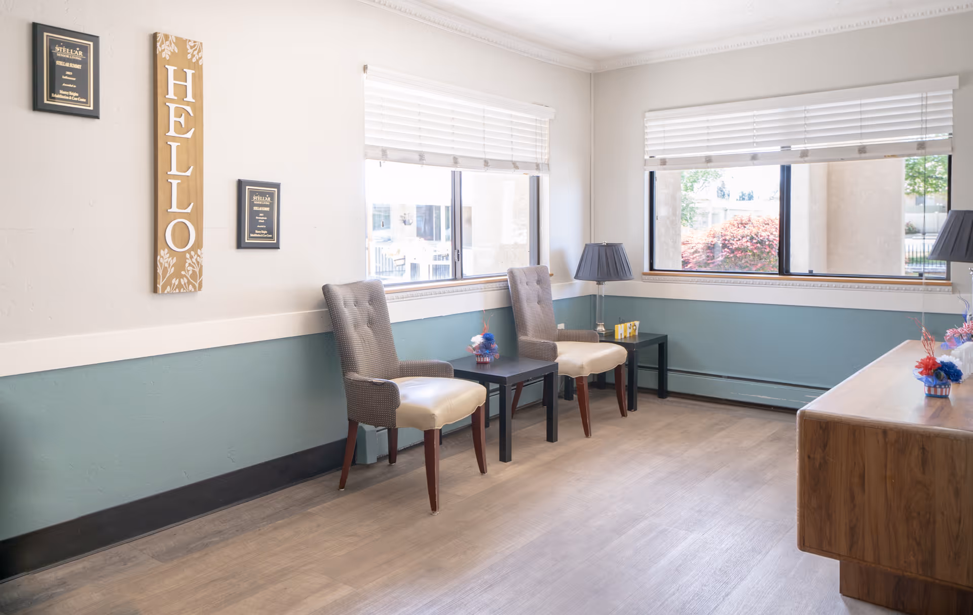 A bright and clean waiting area with two cushioned chairs separated by a small black table, a wooden cabinet on the right, two windows with white blinds letting in natural light, and wall decor including a vertical wooden sign that says 'HELLO' and two framed plaques.