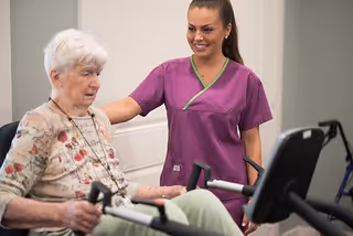 An elderly woman using a stationary exercise bike with the assistance of a smiling caregiver wearing purple scrubs in a light-colored room.