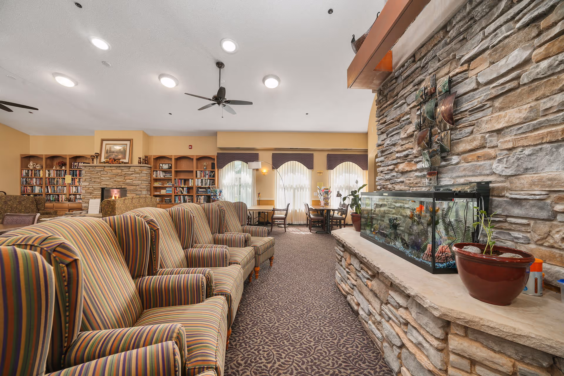 A cozy living room area in The Timbers of Apple Valley featuring a row of striped armchairs, a stone fireplace with a built-in fish tank, a large bookshelf filled with books, and tables with chairs near windows with sheer curtains.