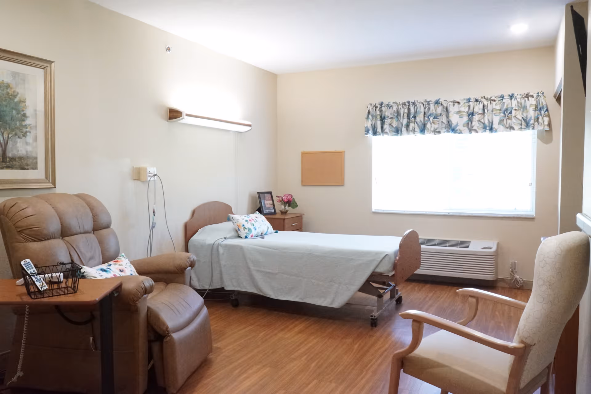 A senior living facility bedroom with a single bed covered in a light blue sheet, a brown recliner chair with a side table holding a remote control and a basket, a wooden armchair, a window with floral curtains, a wall-mounted light, a small wooden nightstand with a framed photo and a flower vase, and a framed painting on the wall.