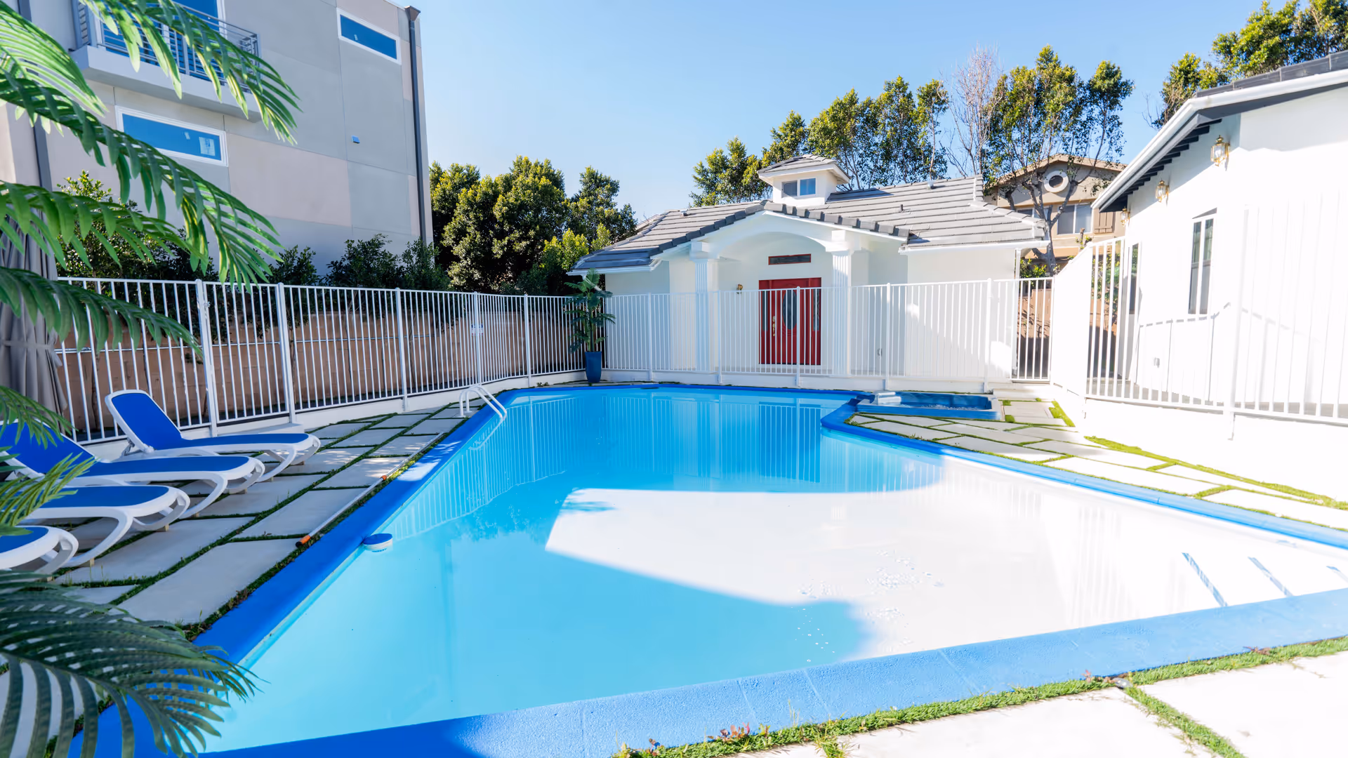 Outdoor swimming pool area with blue water, surrounded by a white fence. There are several blue and white lounge chairs on the left side, and a small white building with a red door in the background. Green plants and trees are visible around the pool area under a clear blue sky.
