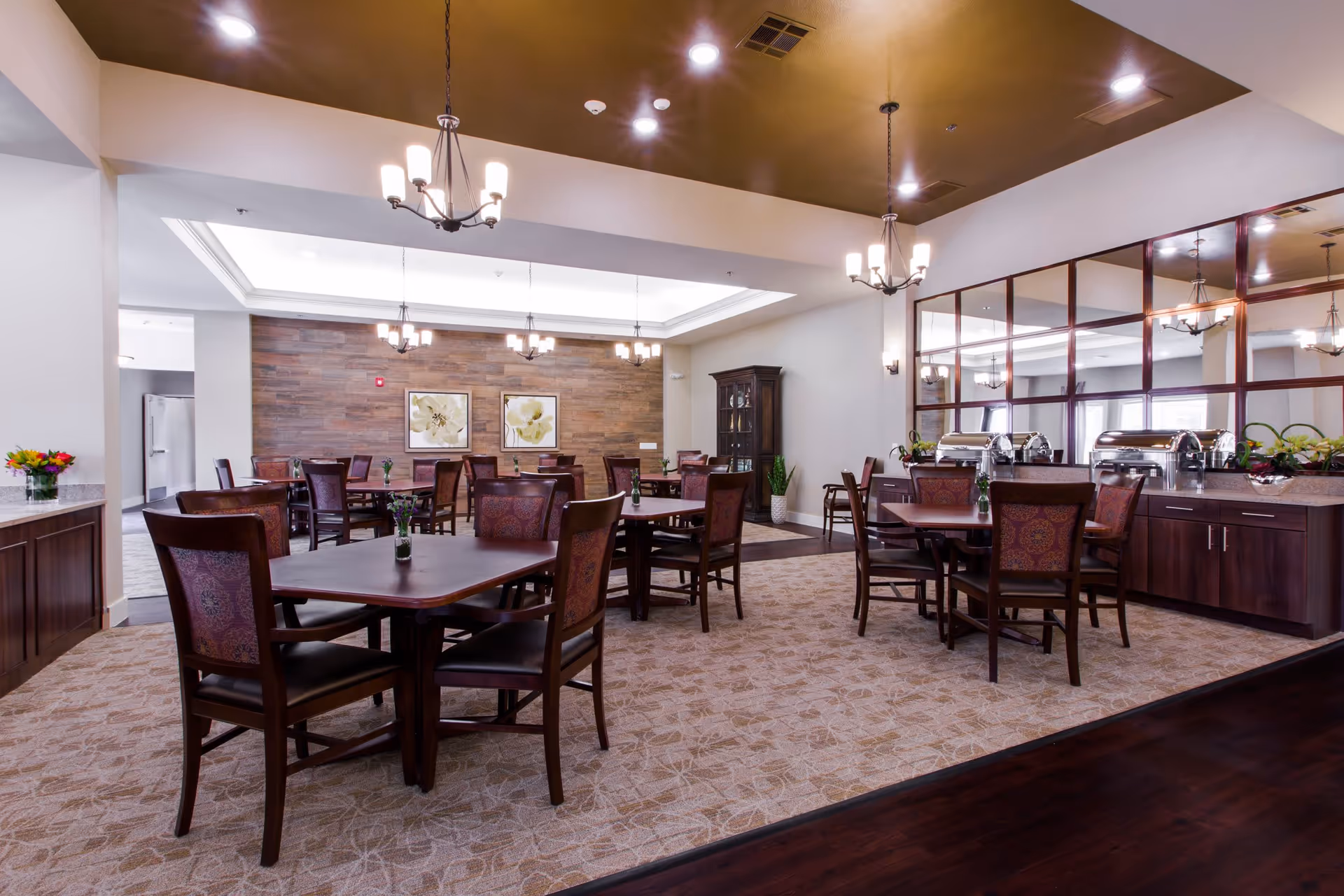 A spacious dining room in an assisted living facility with multiple dark wood tables and chairs arranged neatly. The room features a patterned carpet, a wooden accent wall with two floral paintings, and several chandeliers hanging from the ceiling. There is a buffet area with covered serving dishes and a large mirror on the right side. The overall decor is warm and inviting.