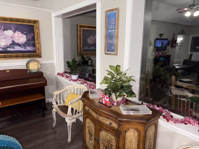 A cozy interior corner of a senior living facility featuring a wooden piano with a decorative plate on top, a vintage-style white chair with a yellow cushion, a wooden cabinet with floral designs, and a large window decorated with a pink garland. A framed floral painting hangs above the piano, and a framed inspirational quote is on the wall next to the window. The room has wood flooring and soft lighting.