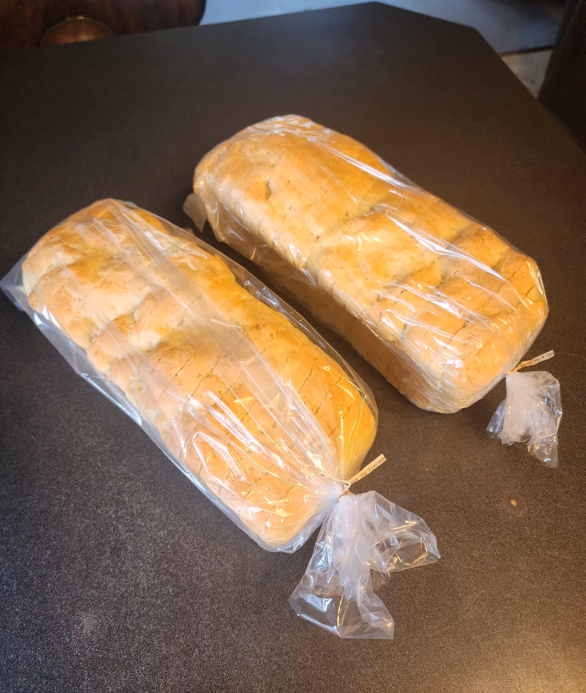 Two loaves of sliced bread wrapped in clear plastic bags placed on a dark countertop.