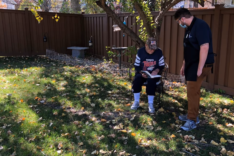 An elderly person wearing a Minnesota Twins sweatshirt and a camouflage cap is sitting on a metal chair reading a book in a fenced backyard. A younger person wearing a face mask, black shirt, and brown pants is standing nearby, looking at the elderly person. The yard has green grass with scattered fallen leaves and a tree providing shade.