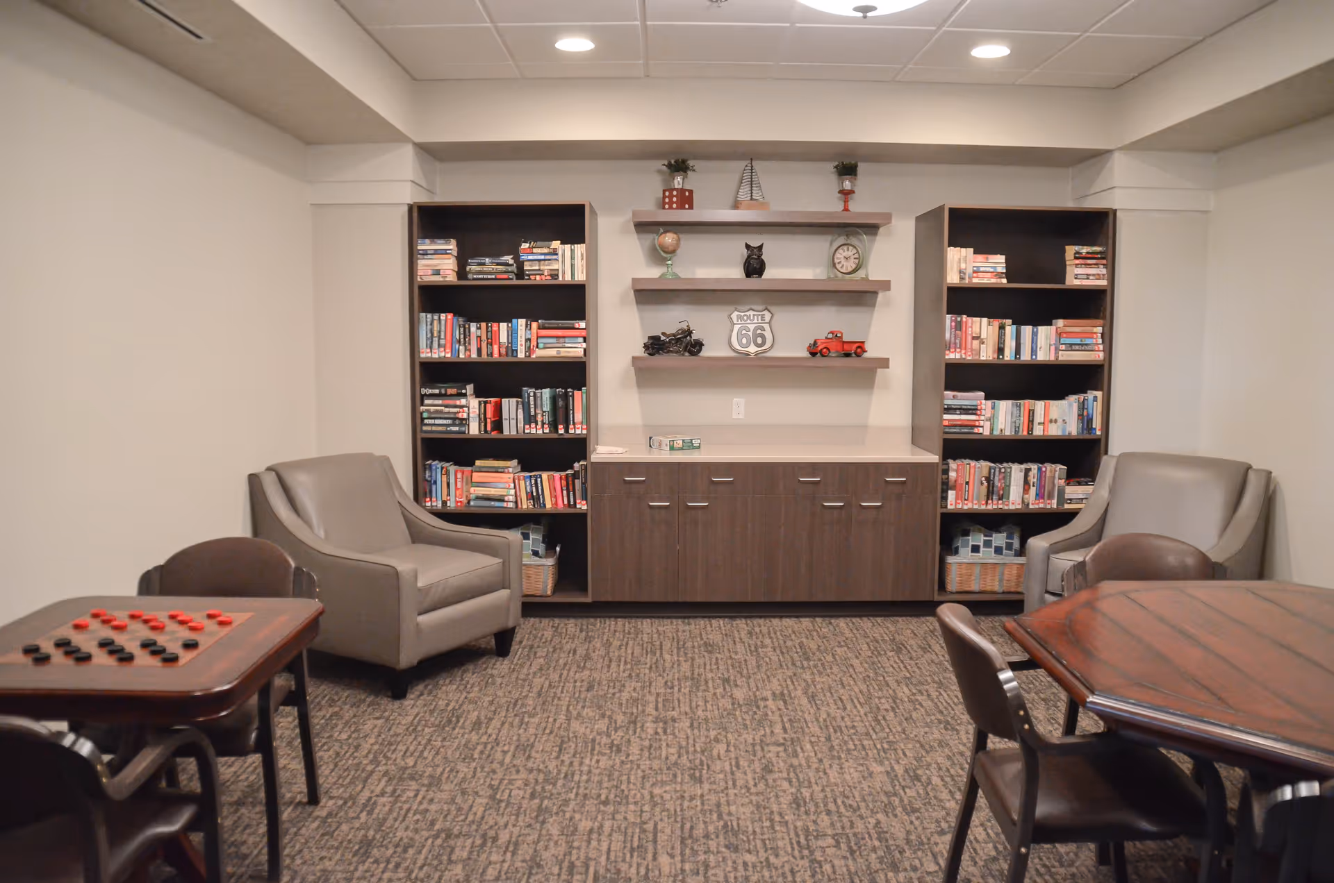 Cozy common room with bookshelves, armchairs, game tables, and decorative shelving.