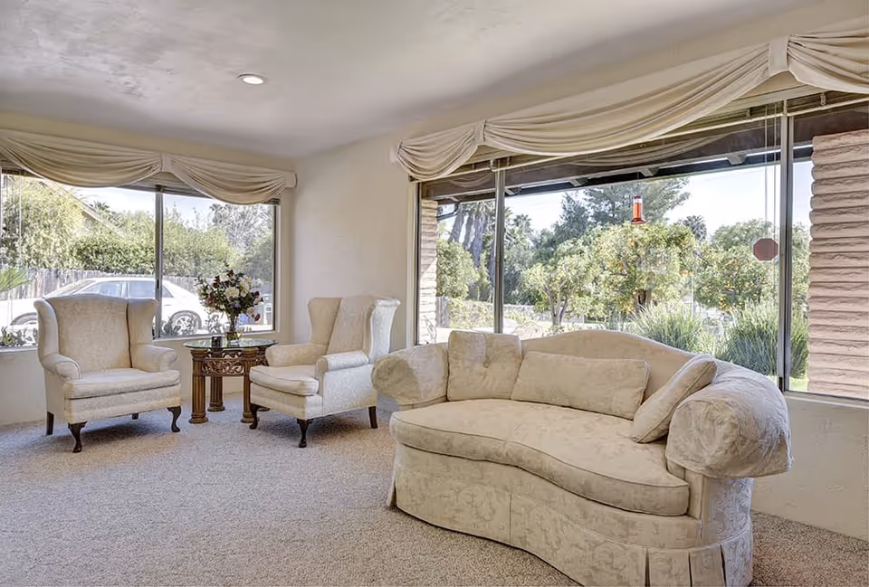 A bright living room with large windows showing a garden outside. The room contains a curved beige sofa with cushions and two matching armchairs around a small wooden table with a flower vase. The walls and carpet are light-colored, and the windows have cream-colored drapes.